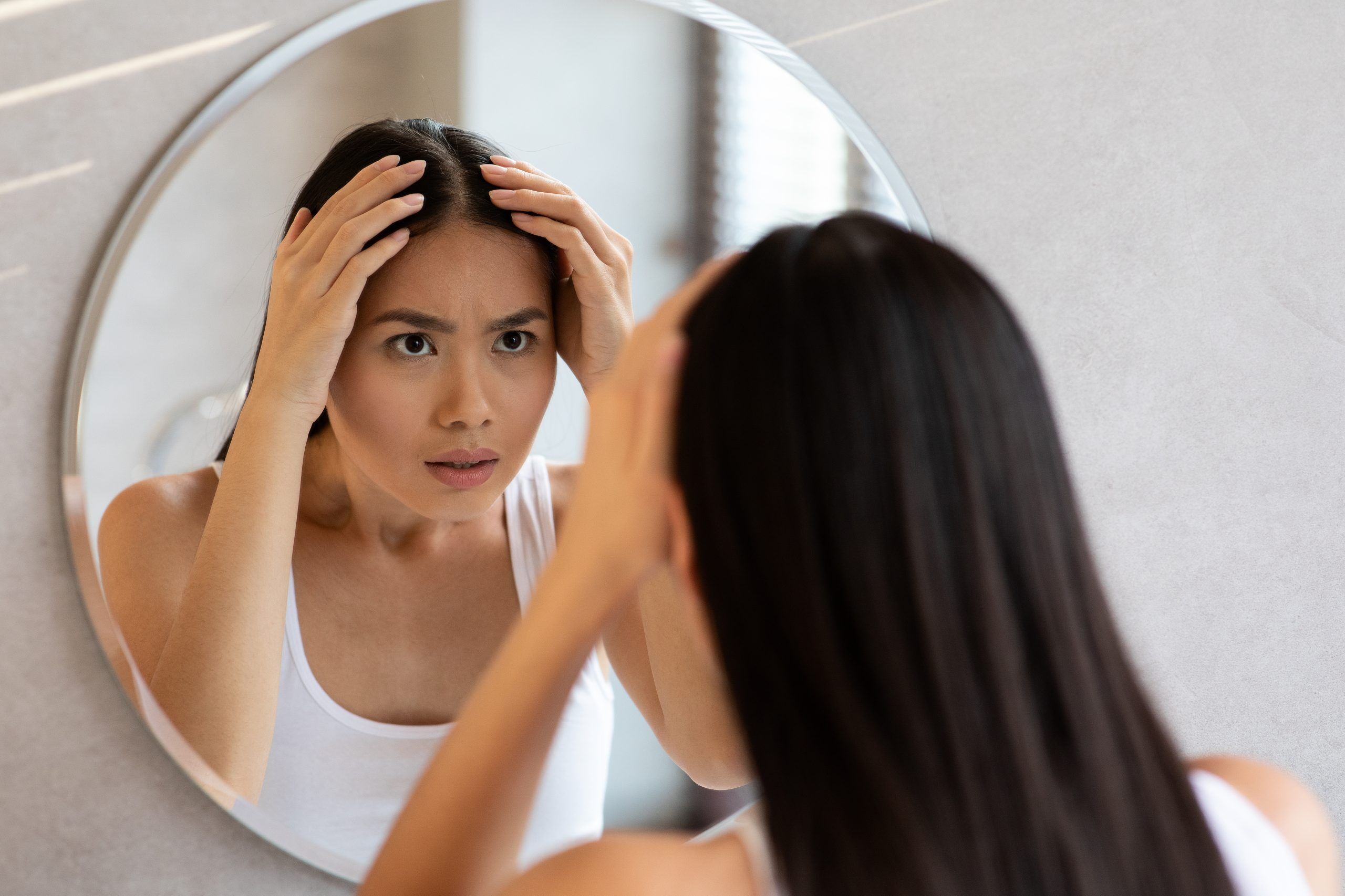 Preocupada mujer de cabello largo mirando el espejo y revisando su cabello, buscando canas, tiro sobre el hombro, interior del baño. Adelgazamiento del cabello, concepto de alopecia